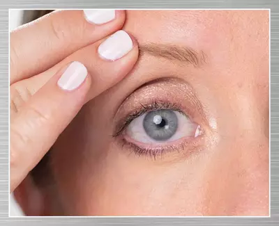A close-up of a woman's eye as she gently lifts her eyelid with one hand, showcasing her eye makeup and a subtle shimmer on her eyelid.
