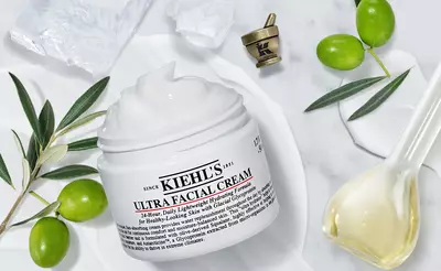 A jar of Kiehl's Ultra Facial Cream is placed on a marble surface, surrounded by green olives, olive leaves, and a small golden measuring scoop. Ice cubes are visible nearby, highlighting a refreshing theme.