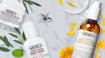 A marble surface featuring Kiehl's skincare products, including Ultra Facial Cream, Ultra Facial Moisturizer, and Calendula Deep Cleansing Foaming Face Wash, surrounded by yellow flowers and green leaves.