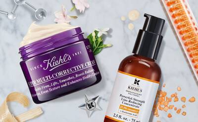 A flat lay of Kiehl's skincare products, featuring a purple jar of Super Multi-Corrective Cream and an orange bottle of Powerful-Strength Line-Reducing Concentrate, surrounded by decorative elements and textures.