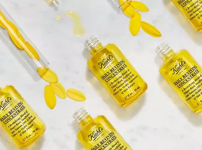 Stekleničke Kiehl's Daily Reviving Concentrate z rumenim oljem in cvetnimi listi na belem marmorju.