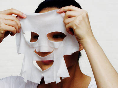 Ženska drži masko za obraz iz papirja pred svojim obrazom v svetli, neutralni okolici.
