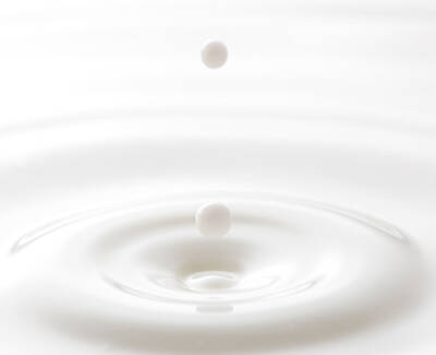 A drop of liquid is about to fall into a smooth, white surface, creating ripples in the water below.