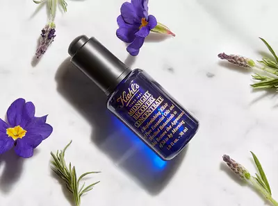 Steklenička modre nočne kreme z rožicami in zelišči okrog, imenovana 'Midnight Recovery Concentrate.'