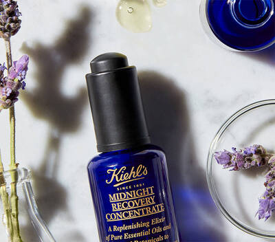 Steklenička Kiehl's Midnight Recovery Concentrate na mramorni površini, obkrožena z sivko in nekaj kapljicami olj.