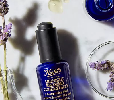 Steklenička Kiehl's Midnight Recovery Concentrate na mramorni površini, obkrožena z sivko in nekaj kapljicami olj.