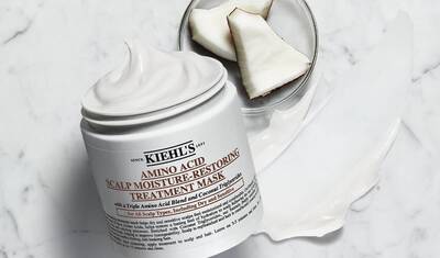 A jar of Kiehl's Amino Acid Scalp Moisture-Restoring Treatment Mask beside a bowl of coconut pieces on a marble surface.